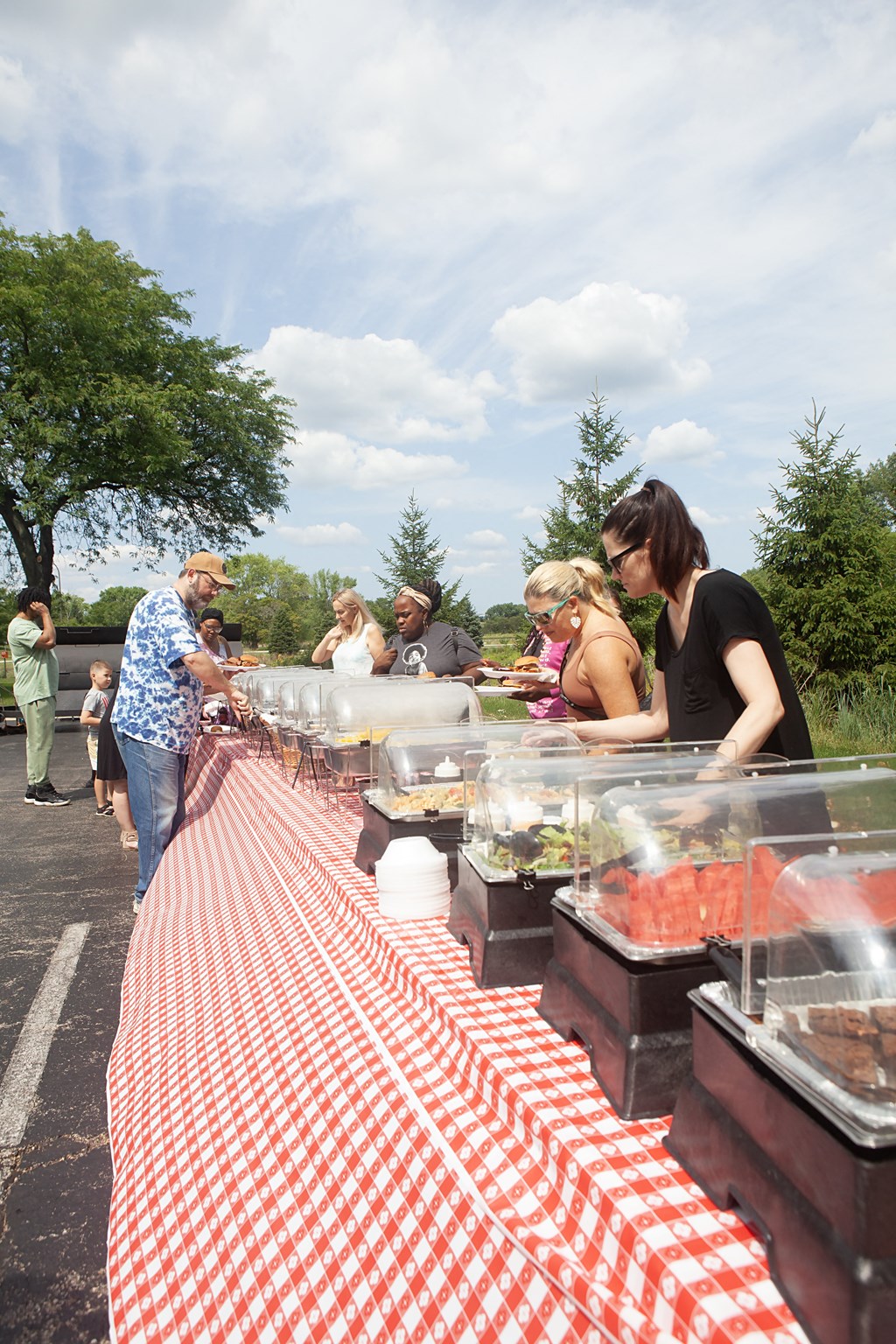 people lined up at an outdoor buffet table with a red and white checkered table cloth