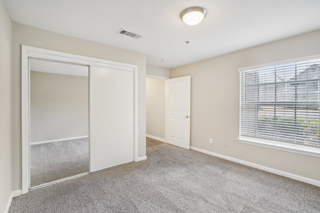 A room with a carpeted floor, a window with blinds, and a door leading to another room.