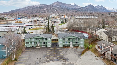 A parking lot with two green buildings in the foreground and mountains in the background.