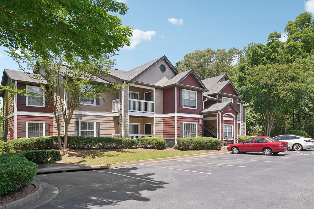 A red car is parked in a parking lot in front of a two-story apartment building.