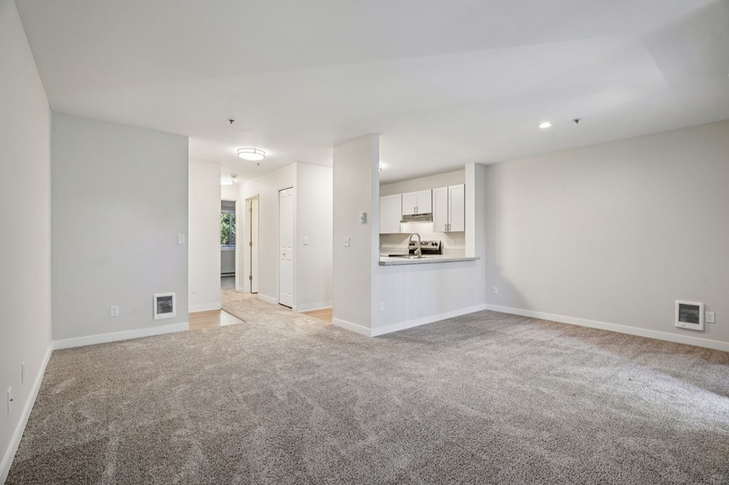 A spacious room with a carpeted floor and a kitchen area in the background.
