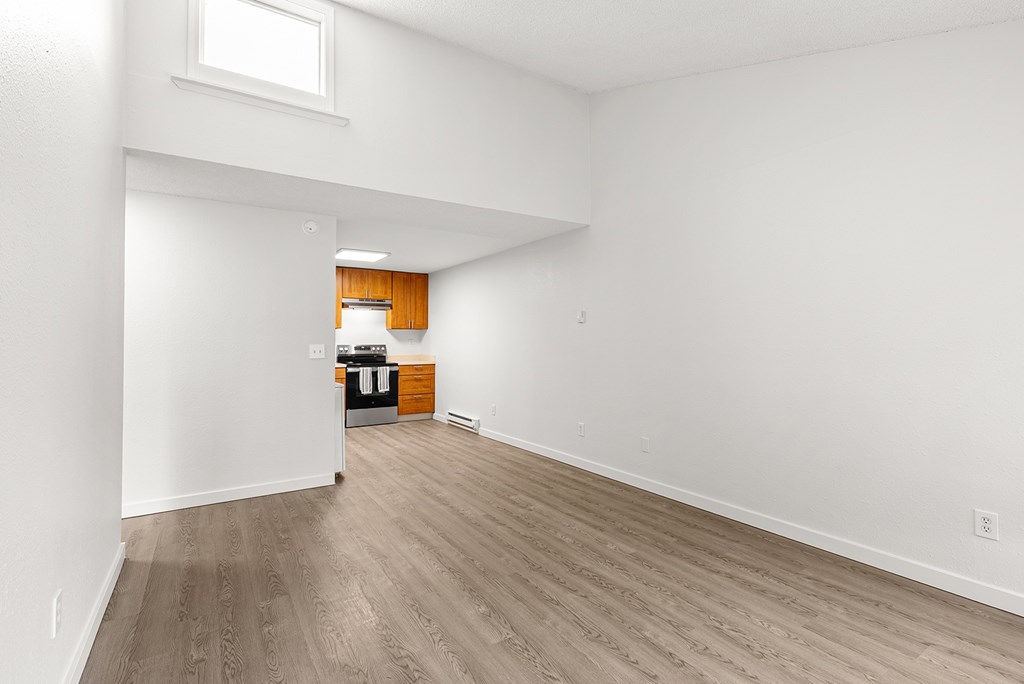 A room with wooden flooring and white walls and kitchen in the back