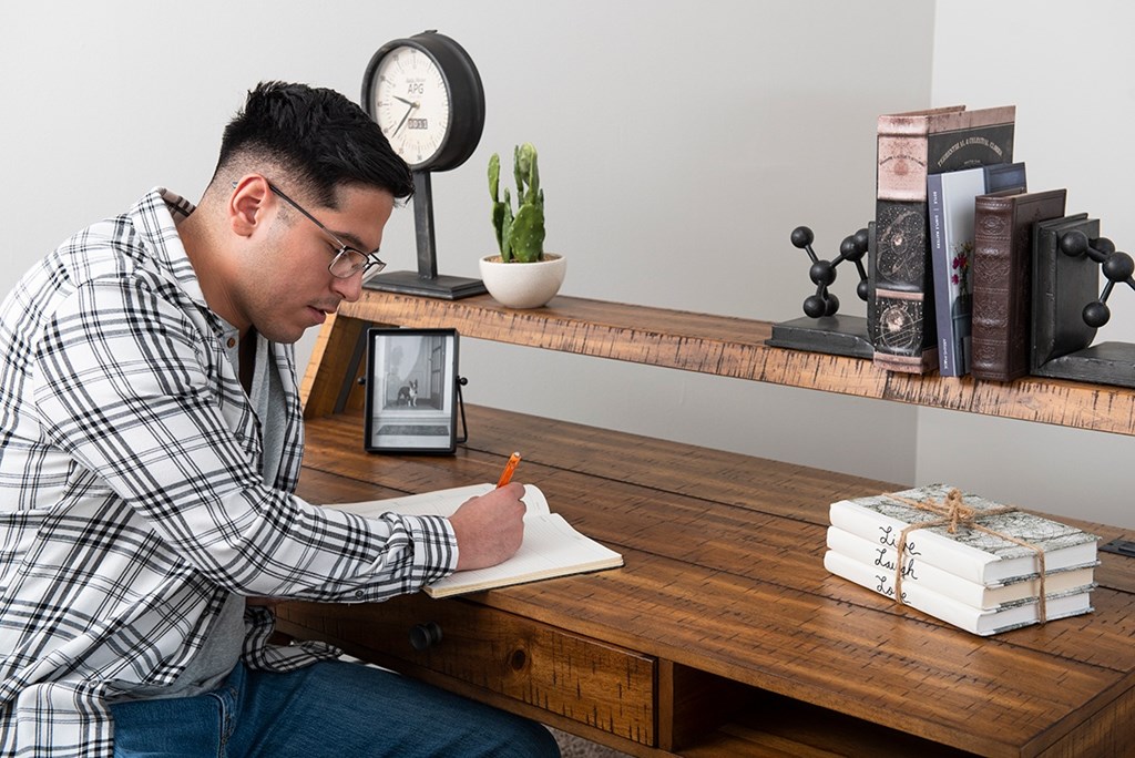 A man is writing on a piece of paper while sitting at a wooden desk with a clock, books, and a small potted plant on the shelf behind him.