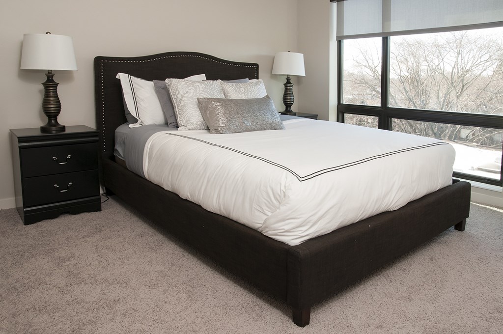 A bed with a white sheet and pillows is in a room with a carpet and two lamps.