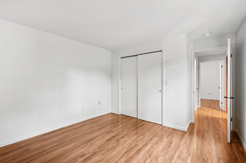 A room with white walls and wood flooring with a white door on the left and a white closet with a red handle on the right.