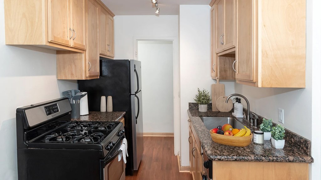 A kitchen with a black stove and refrigerator, wooden cabinets, and a granite countertop with a bowl of fruit on it.
