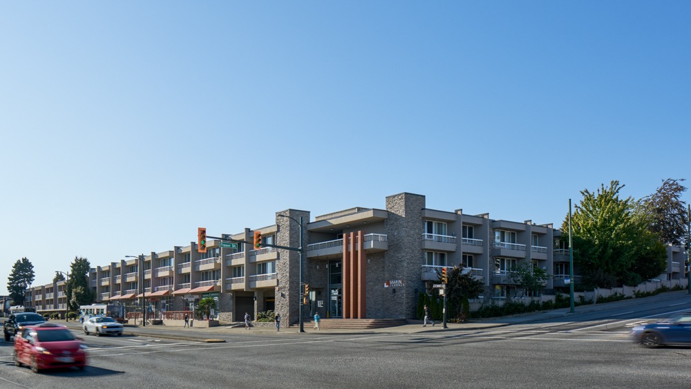 A red car is driving on a street past apartment buildings.