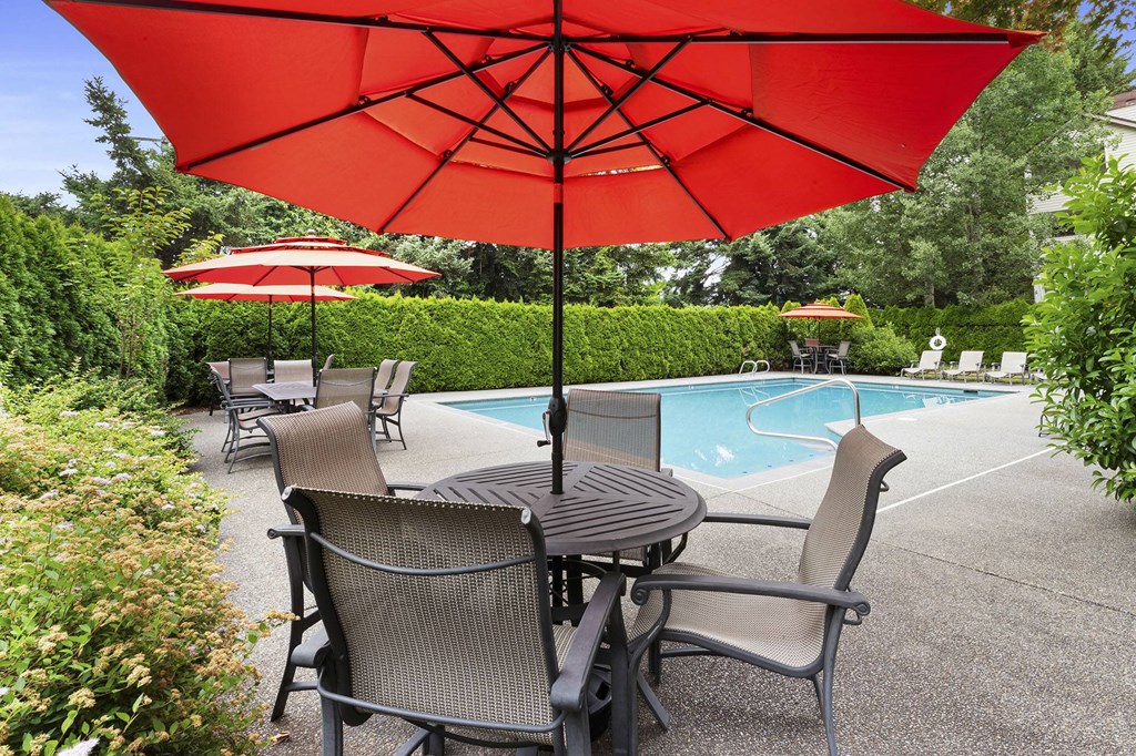 A red umbrella is over a table with chairs by a pool.