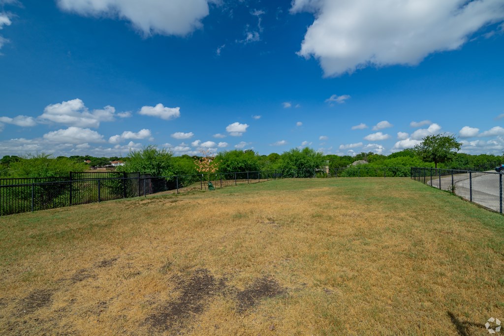 A large, open field with a fence and trees in the distance under a blue sky with clouds.