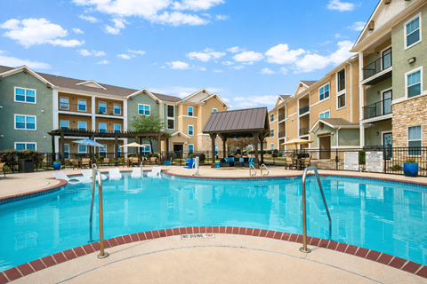 Apartments for Rent in Converse, TX - The Heights at Converse - Resort-Style Swimming Pool With Lounge Seating, Covered Cabana, Wooden Pergolas, Metal Fence, and Apartment Buildings.