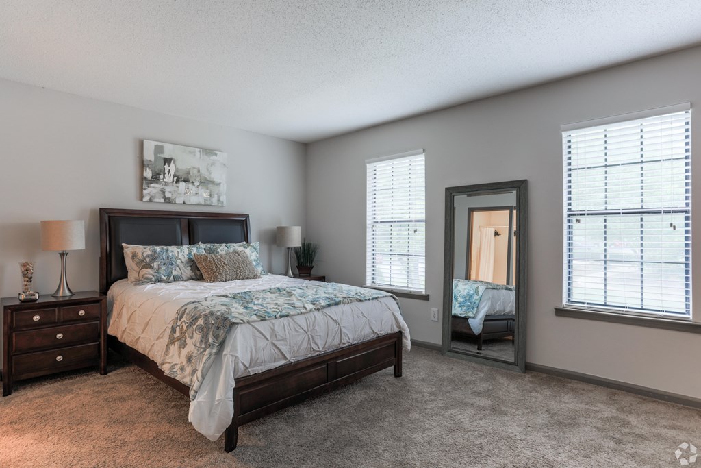 A bedroom with a large bed, a nightstand with a lamp, and a window with blinds.