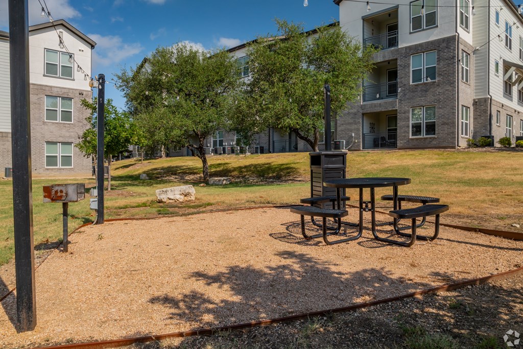 A picnic table sits in the middle of a gravel area with apartment buildings in the background.