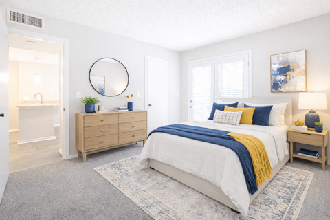 2-Bedroom Apartments in Duluth, GA - The Eclipse - Bedroom With King Bed, Blue And Yellow Accent Pillows, Nightstand, Dresser, Area Rug, And Large Windows.