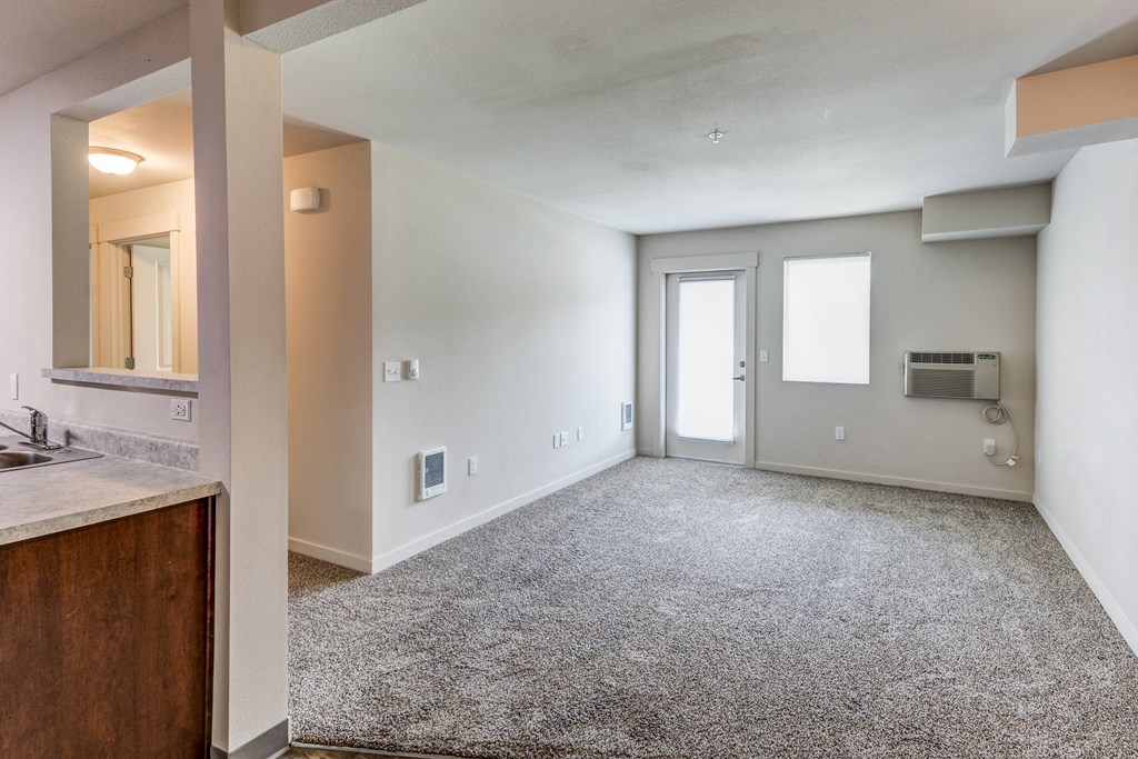 an empty living room with a kitchen and a door to a hallway