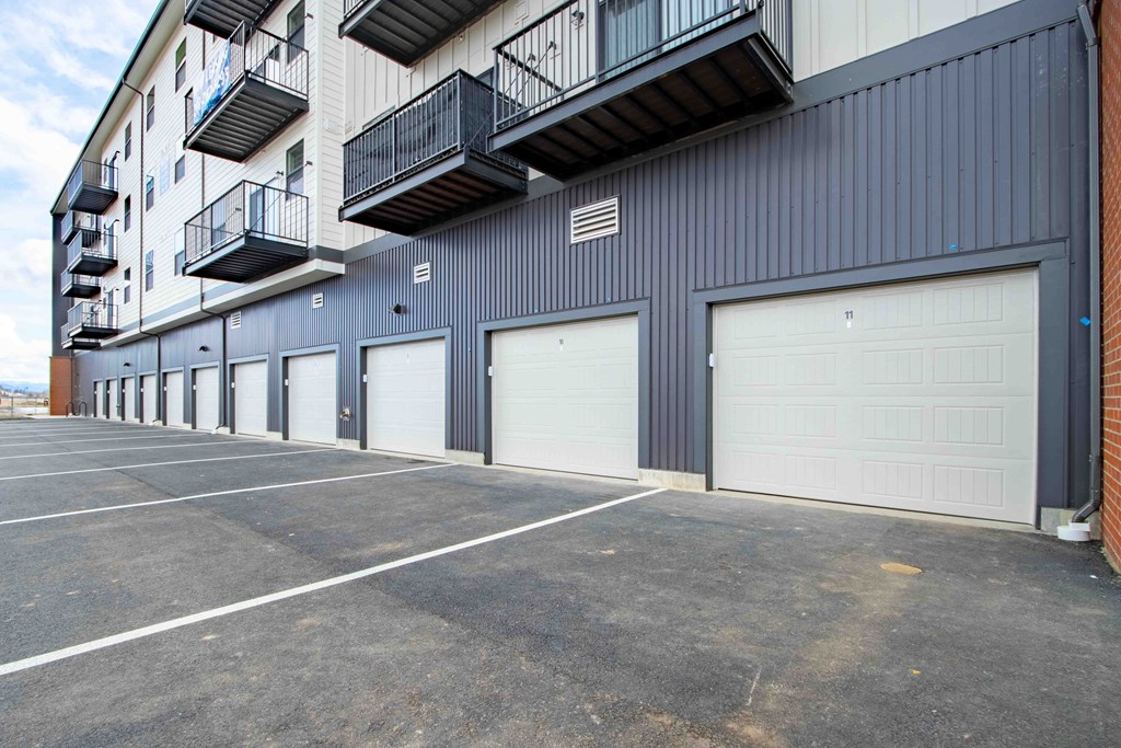 A parking lot in front of a grey building with a balcony on the second floor.