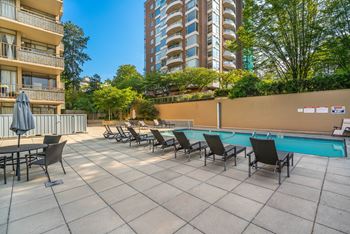 A pool area with chairs and a table is surrounded by buildings and trees.