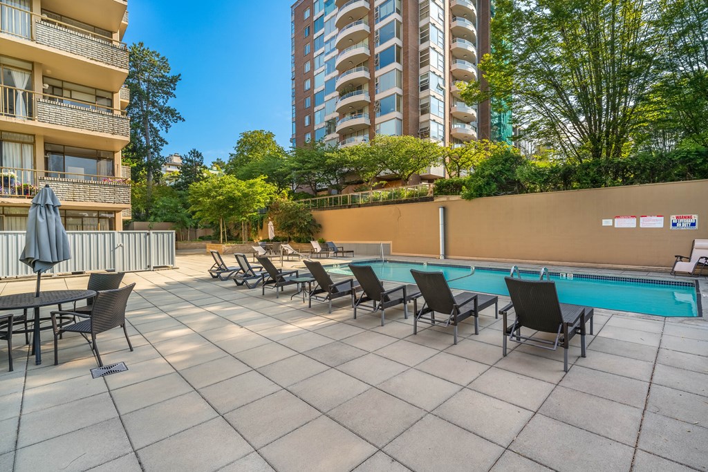 A pool area with chairs and a table is surrounded by buildings and trees.