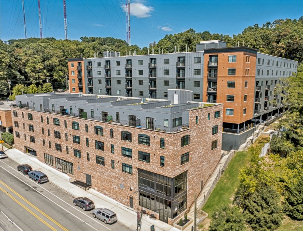 A large multi-story building with a mix of brick and modern architecture sits on a street corner with cars parked along the side.