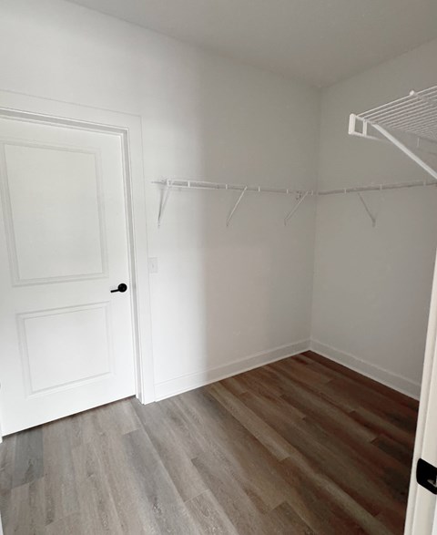 A white door is on the left of a room with a white wall and a wooden floor.