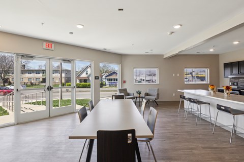 A large room with a long table and chairs, a kitchen area, and a view of the outside through the glass doors.