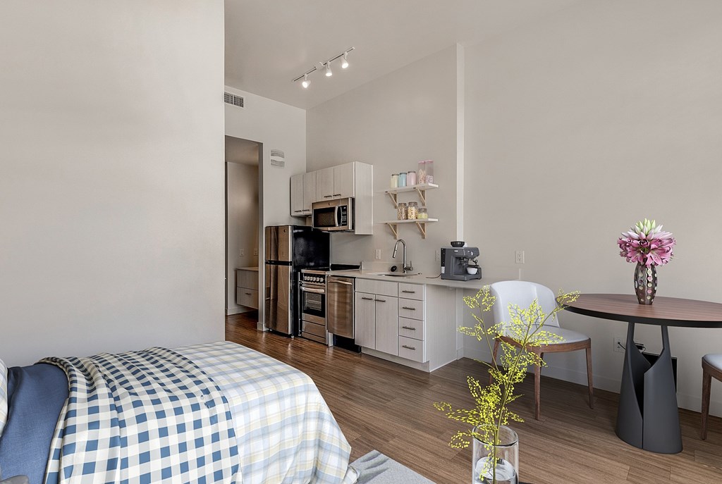 A bedroom with a bed, a table with a vase of flowers, and a kitchen area with appliances. at RiDE at RiNo Apartments, Denver, CO, 80216