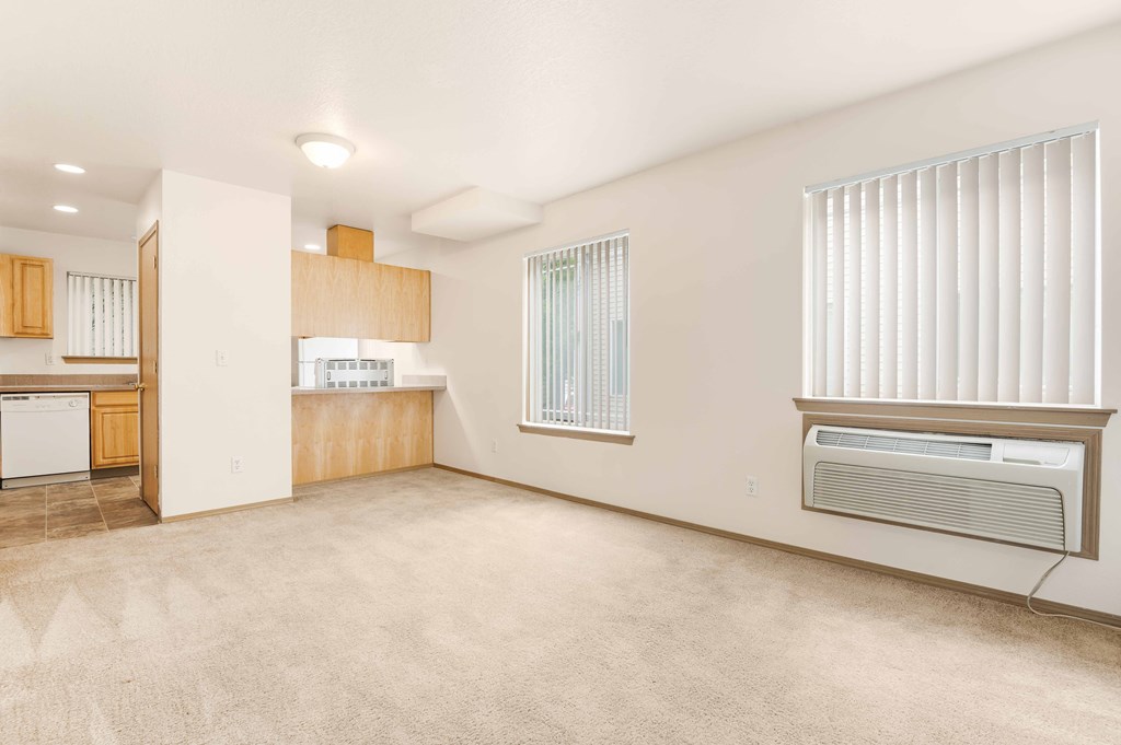 an empty living room with two windows and a kitchen in the background