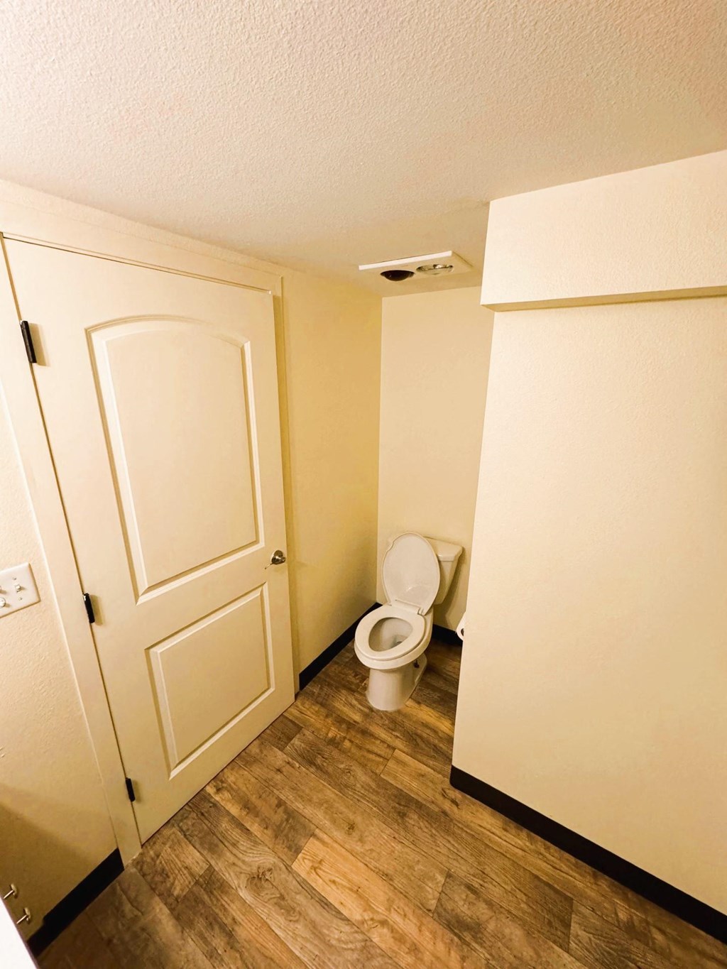 A white toilet sits in a small bathroom with wood floors and a white door.