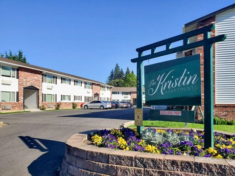 The entrance to The Kristin Apartment complex with a sign and flowers.