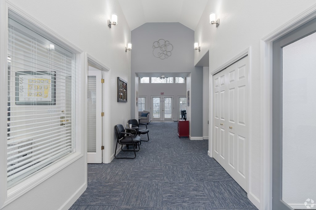 A hallway with a carpeted floor, white walls, and a row of chairs.