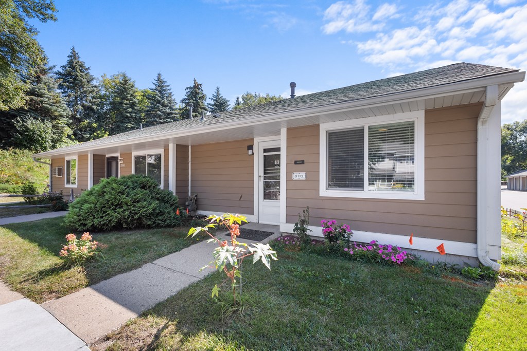 A small house with a brown and white exterior is surrounded by greenery and flowers.