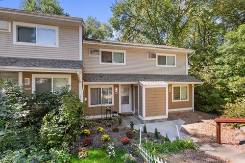 A house with a brown and beige exterior is surrounded by greenery and a garden with flowers.