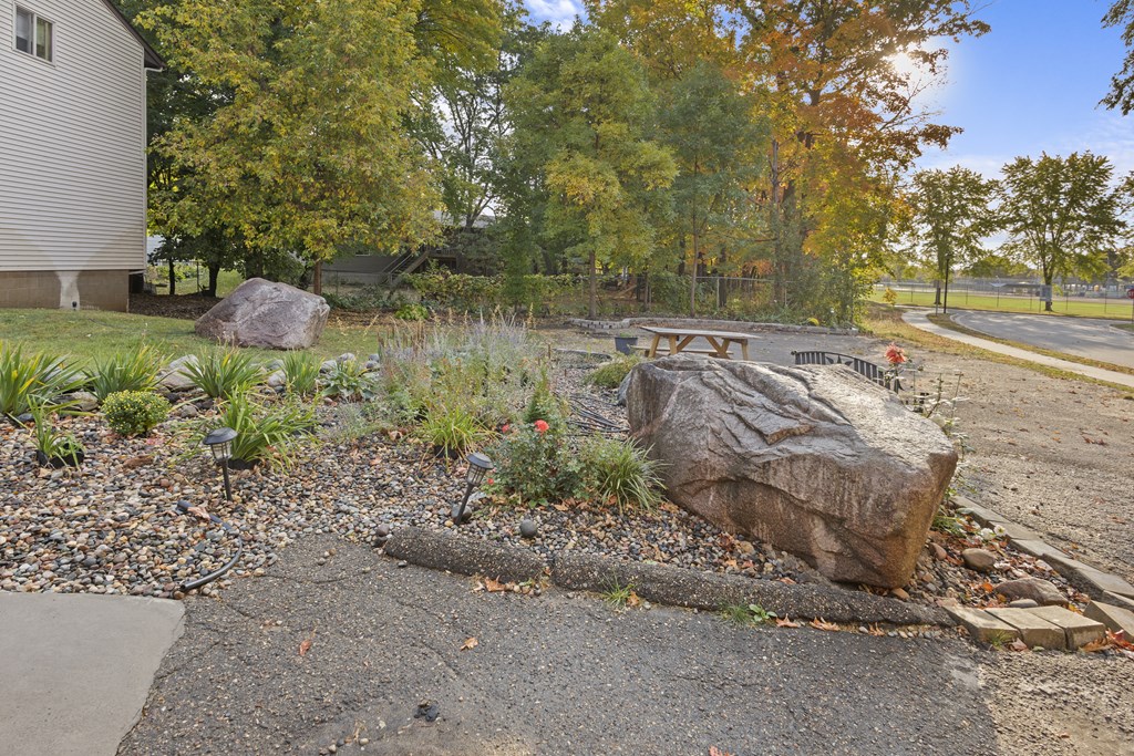 A large rock sits in the middle of a gravel area with a grassy area and trees in the background.