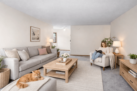A woman is sitting on a couch in a living room with a cat lying on the floor.
