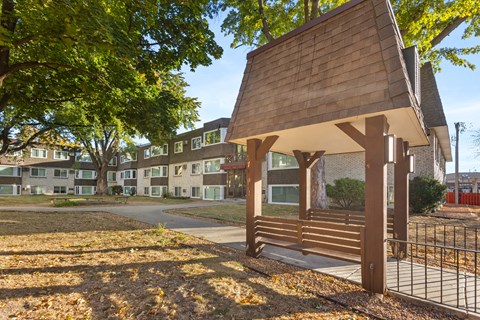 A wooden pavilion sits in the middle of a grassy area with a fence and apartment buildings in the background.