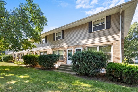 A house with a brown siding and a white door is surrounded by green bushes and trees.
