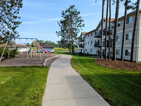 A playground with a slide and swings is surrounded by a grassy area and a sidewalk.