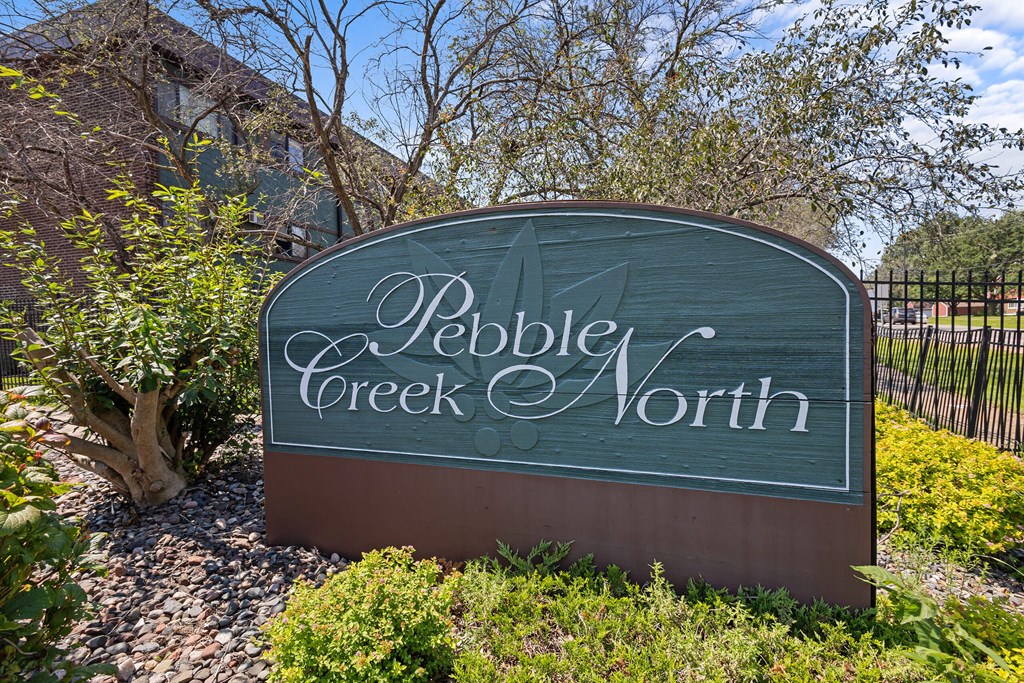 A sign that says Pebble Creek North is shown in front of a fence and some trees.