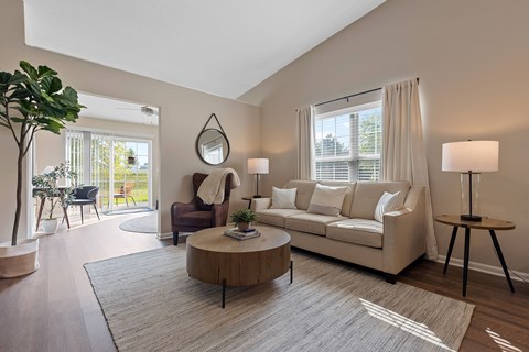 A living room with a white couch, a wooden coffee table, and a large window with white curtains.