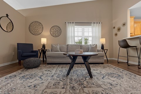 A living room with a grey sofa, a black armchair, a round ottoman, a table with a lamp and a rug.