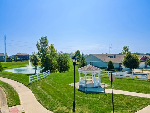 A gazebo sits in the middle of a grassy area with a fence and houses in the background.
