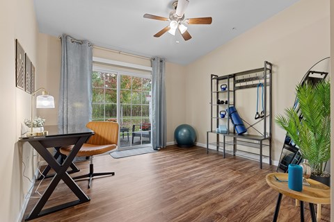 A room with a wooden floor, a ceiling fan, a table with a blue bottle, a chair, a window with a view of trees, a gym equipment, and a potted plant.