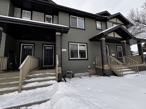 A house with a grey exterior and a black door is shown with snow on the ground.