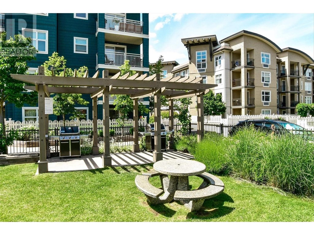 A wooden pergola with a table and chairs is in the foreground of a grassy area with apartment buildings in the background.