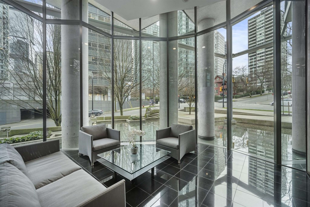 A glass table is surrounded by grey couches in a room with large windows.