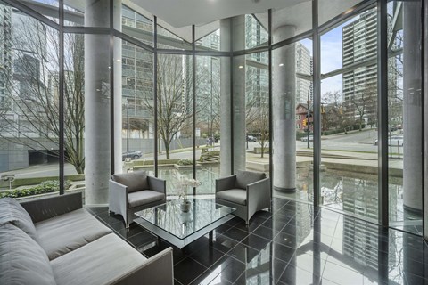 A glass table is surrounded by grey couches in a room with large windows.