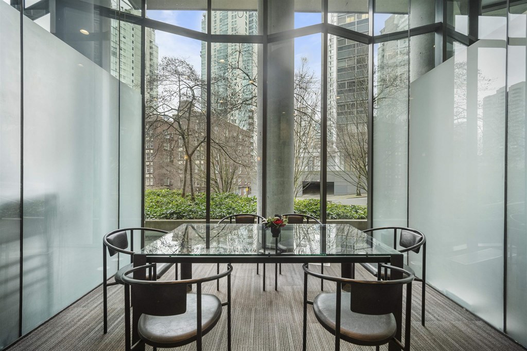 A glass table with chairs in front of a window with a view of trees and buildings outside.