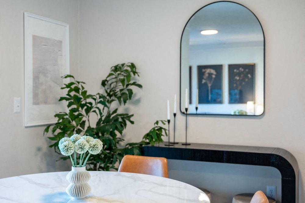 A white vase with white flowers sits on a table in front of a mirror.