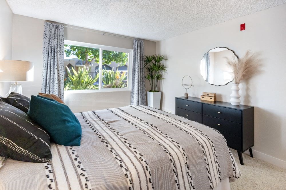 A bedroom with a bed, nightstand, and a window with plants outside.