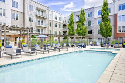 A swimming pool surrounded by lounge chairs and trees in front of apartment buildings.