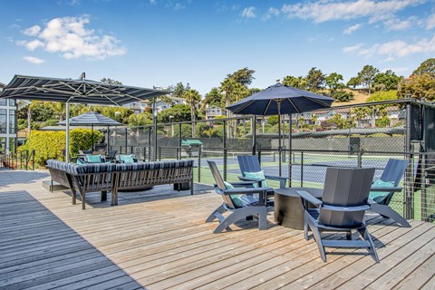 A wooden deck with chairs and umbrellas overlooking a tennis court.